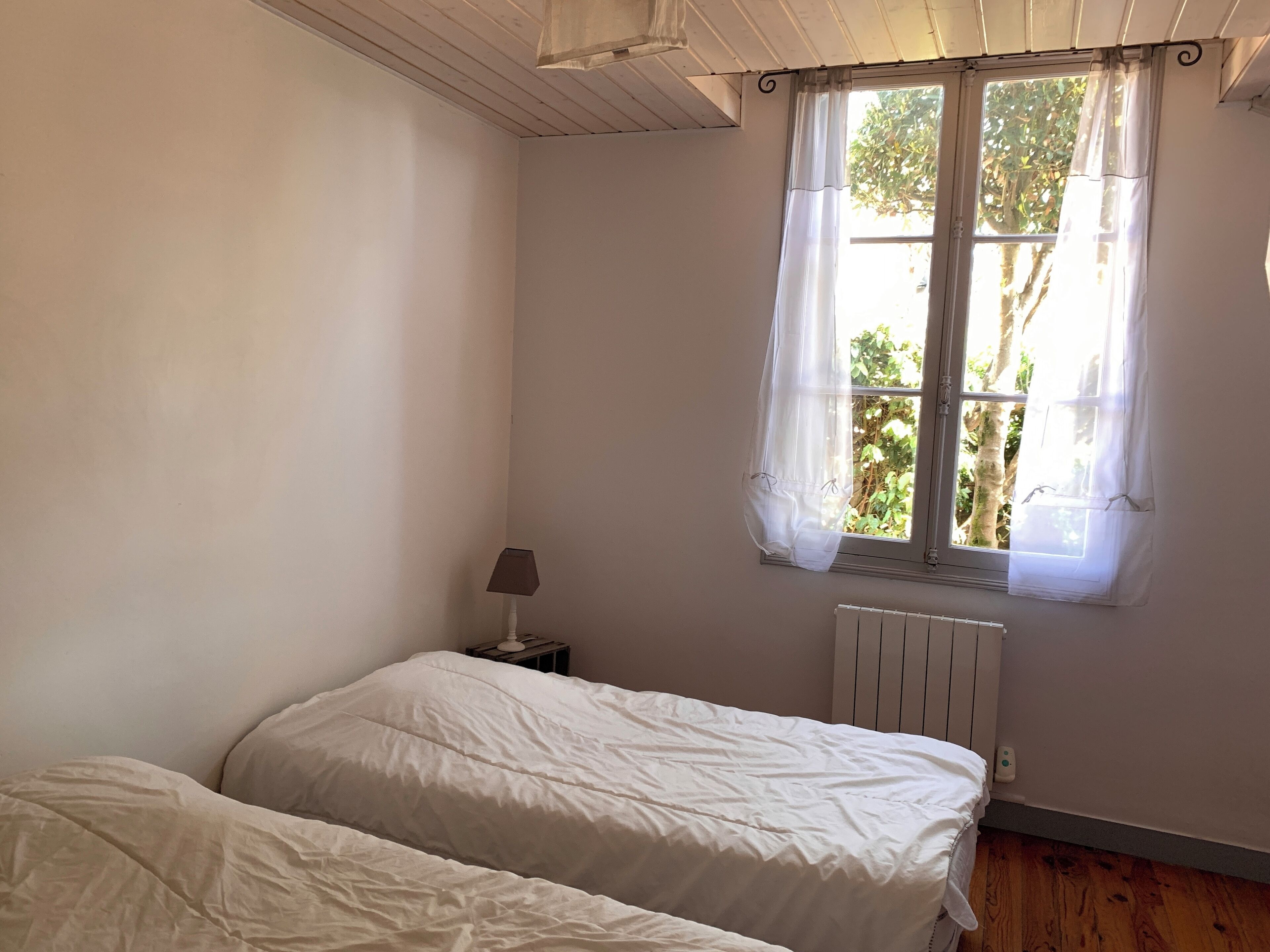 Et soveværelse med to senge, et natbord med lampe, et vindue med let gardin og en radiator.