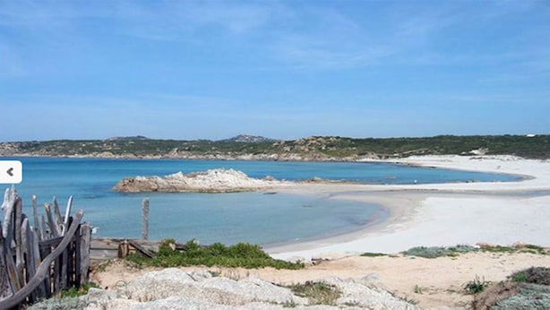 A coastal landscape with a sandy beach, a clear blue sea, and rocky outcrops.