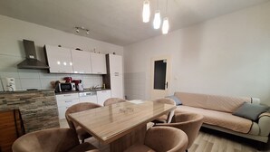 A modern kitchen with white cabinets, a built-in microwave, and a stove. There is a dining area with a wooden table and four chairs. A beige sofa is against the wall, and a door is visible in the background. - App with jacuzzi, fitness near old town (Trogir)