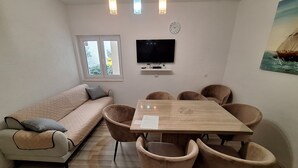 A small, cozy room with a sofa, a wooden dining table with chairs, a flat-screen TV mounted on the wall, and a window with a view of trees. - App with jacuzzi, fitness near old town (Trogir)