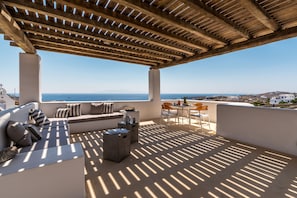 Área de estar ao ar livre com iluminação natural e vista para o mar, com pérgola de madeira, sofá e uma mesa pequena.