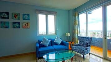 Un salón con sofá azul, una mesa de centro de cristal y un sillón estampado. Hay dos ventanas con marcos blancos y vistas al exterior.