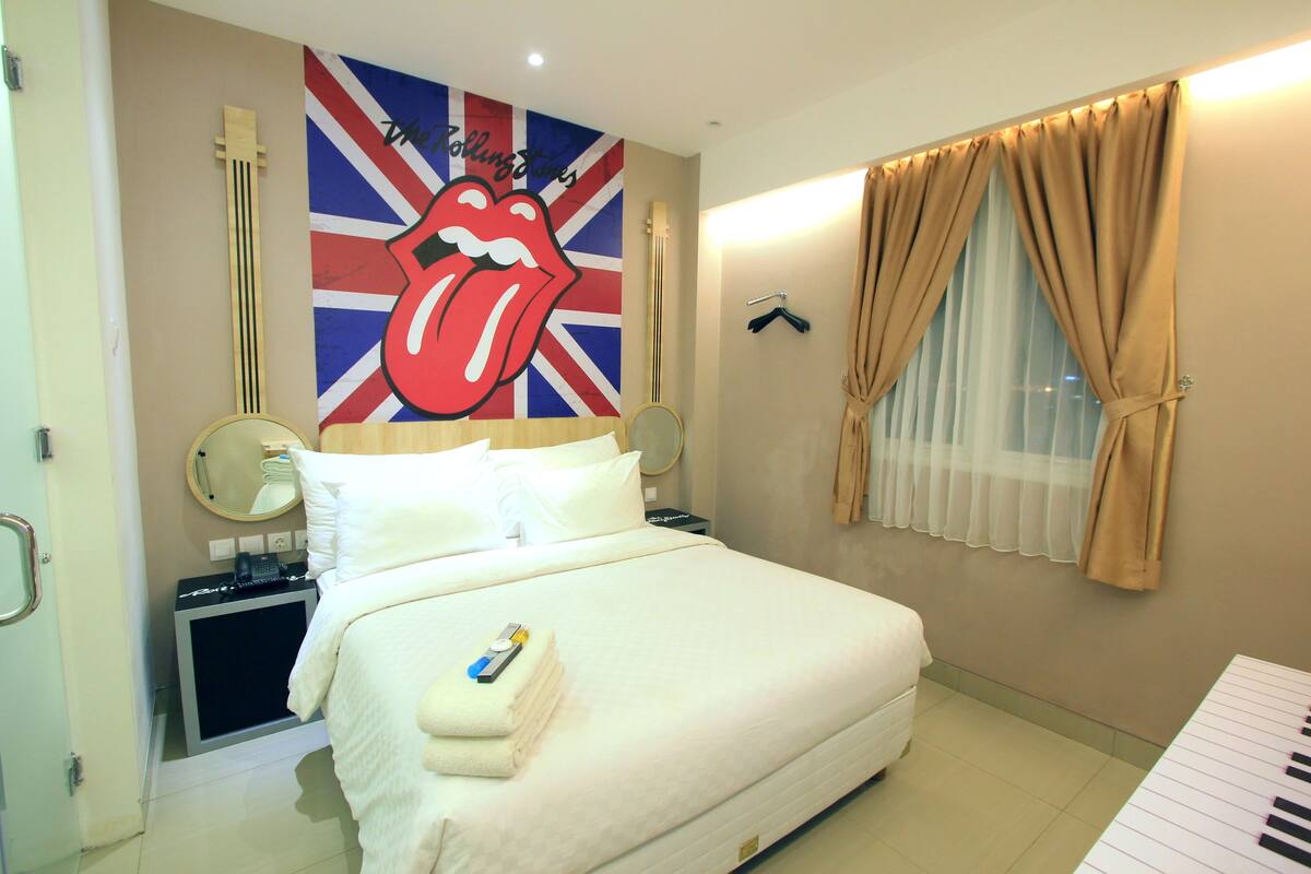 Una habitación de hotel con cama, espejo, guitarra, teclado y un cartel de Rolling Stones.