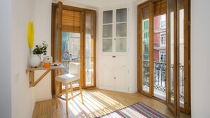 A corner room with a wooden floor, a white chair, a small table with a yellow vase and a plant, and a window with wooden blinds. - Singular Stays El Balcon de Manet (Valencia / València)