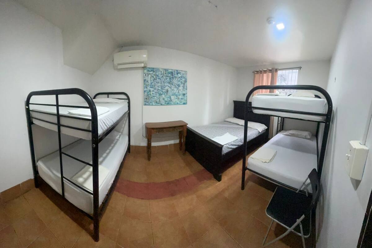 A dormitory room with bunk beds, a small table, a bench, and an air conditioning unit.