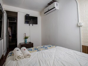 A hotel room with a bed, a TV mounted on the wall, an air conditioning unit, a small table with a vase, and a door leading to another room. - Hostel the pod 33 (Panama City)