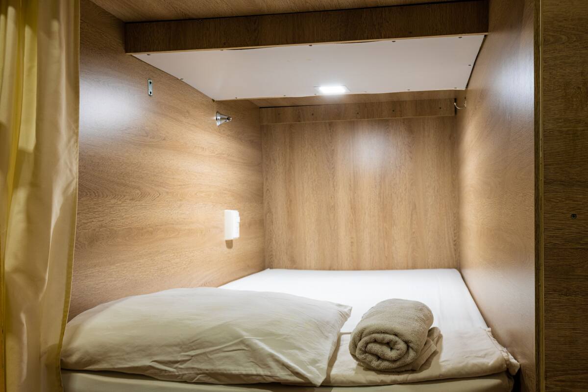 A small, wooden-paneled room with a single bed, a white pillow, and a rolled towel.