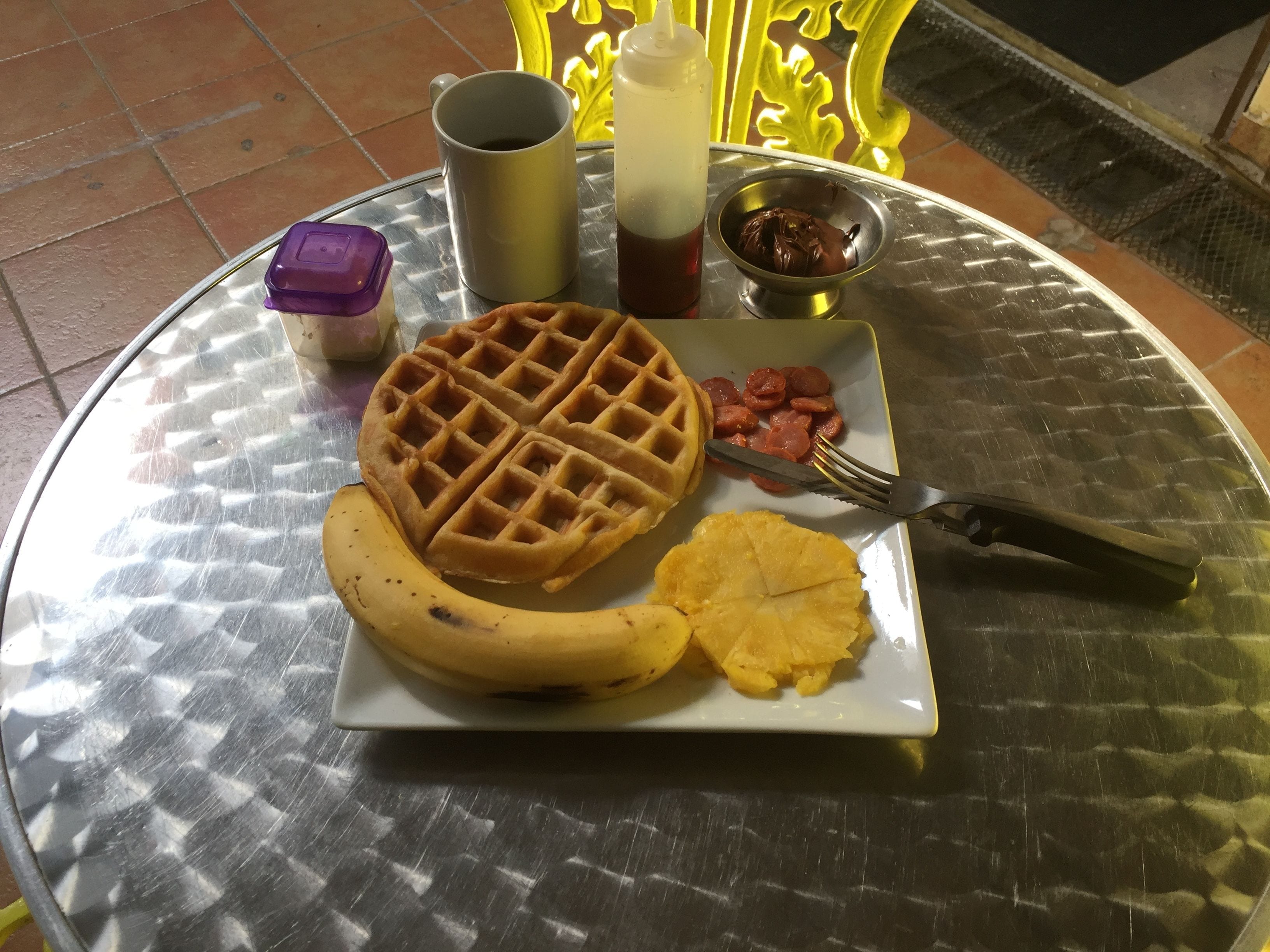Une assiette de petit-déjeuner comprenant une gaufre, une banane, un ananas et une saucisse, accompagnée d’une tasse de café et de condiments, le tout posé sur une table en métal.