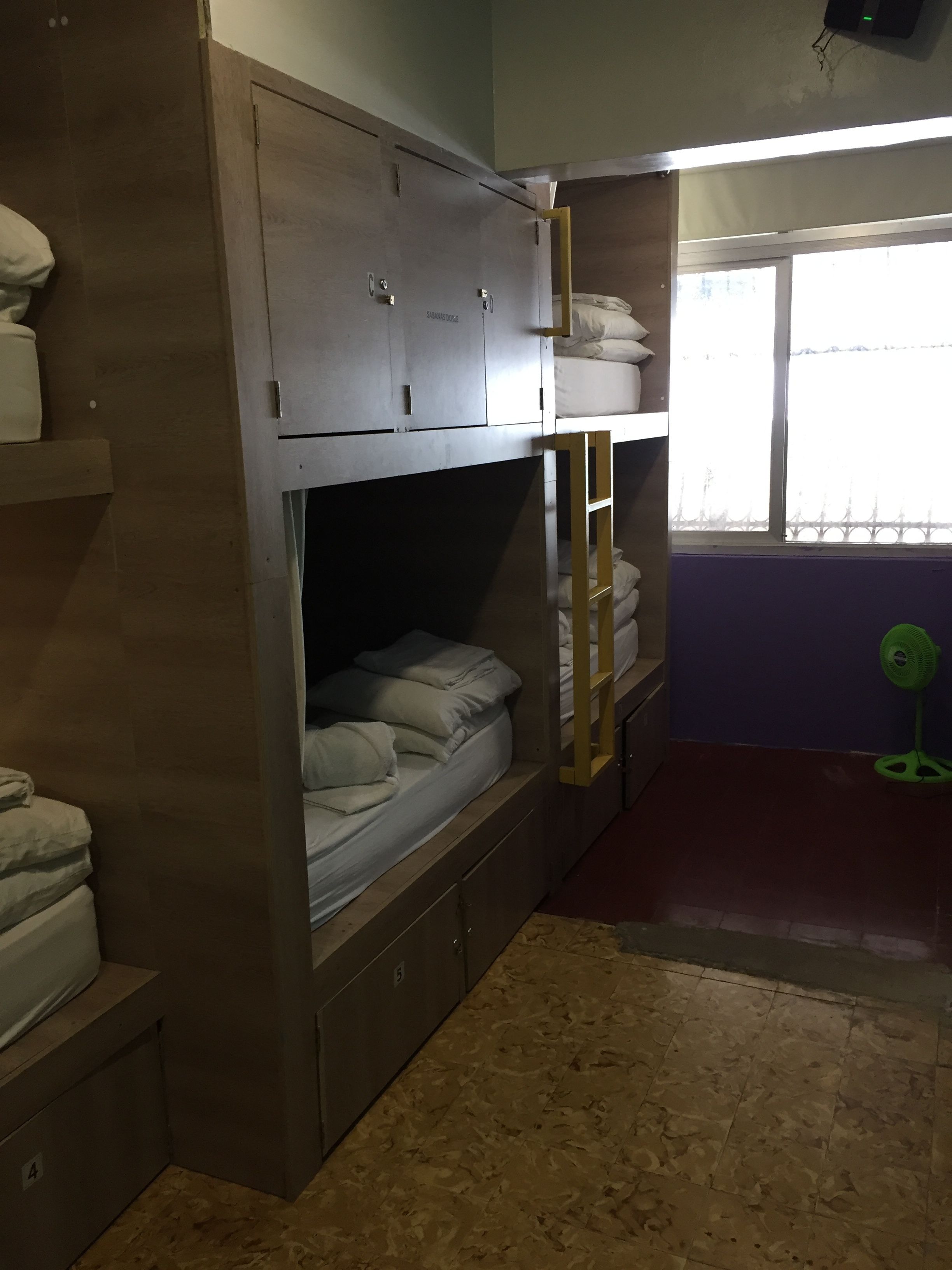A dormitory room with bunk beds, a window with a view, and a green fan.
