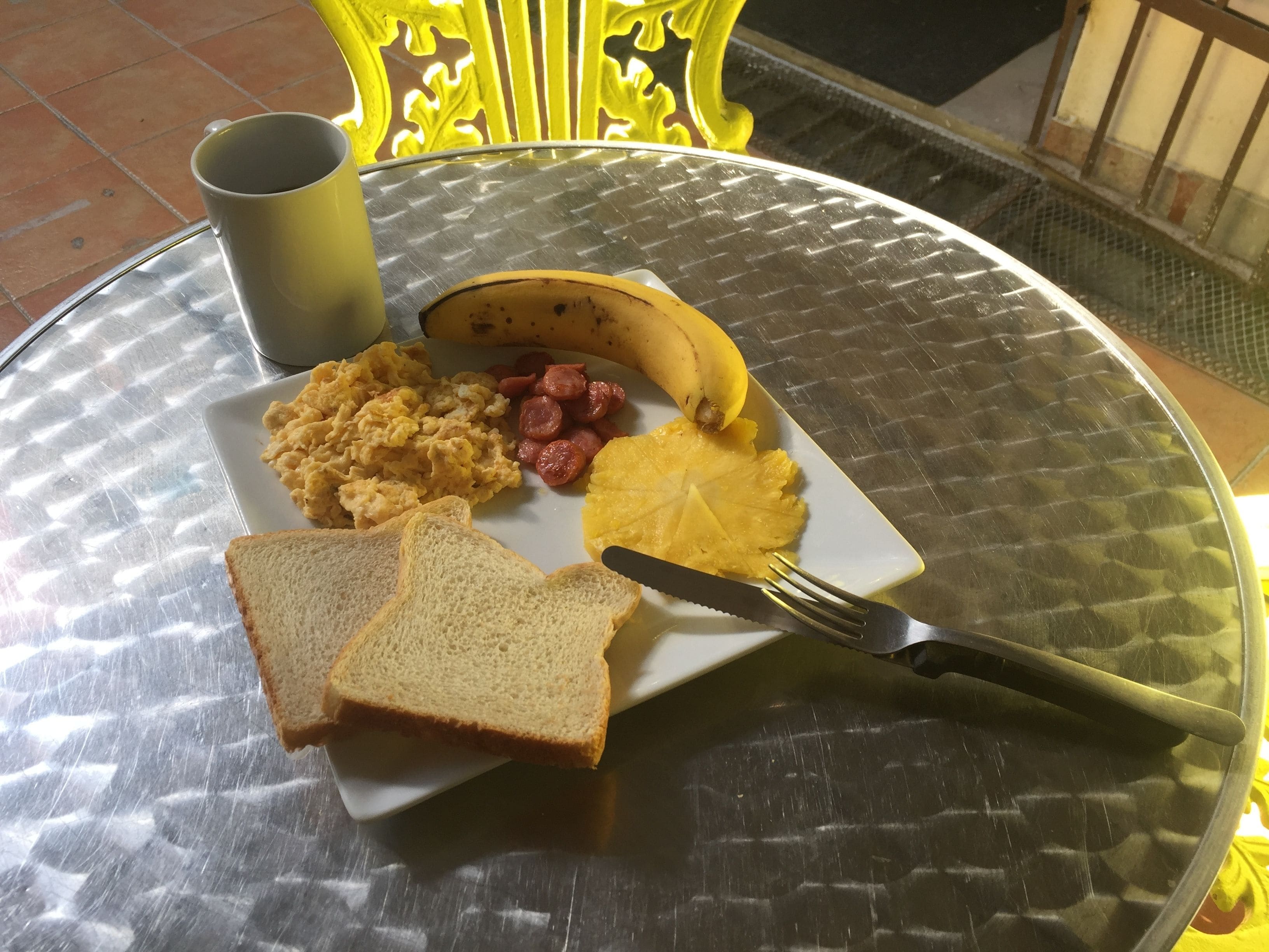 A breakfast plate with scrambled eggs, sausage, a banana, pineapple, and toast on a glass table.