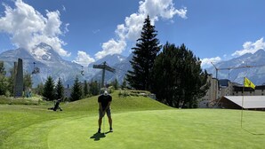 Golfkenttä, jonka taustalla kohoavat vuoret, sekä hiihtohissi ja lipputanko.