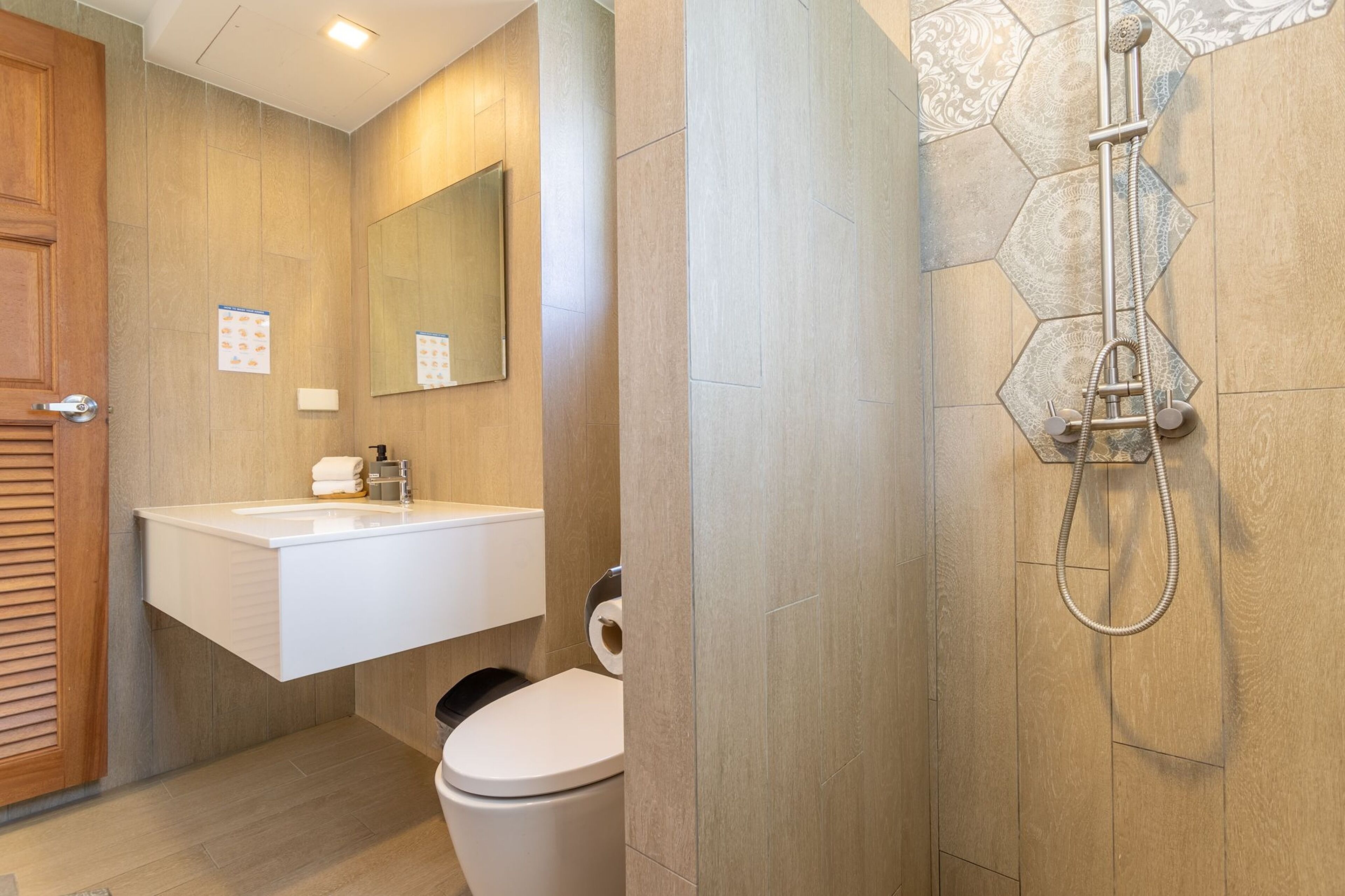 A modern bathroom with a wooden door, a white sink with a mirror, a toilet, and a shower with a hexagonal tile pattern.