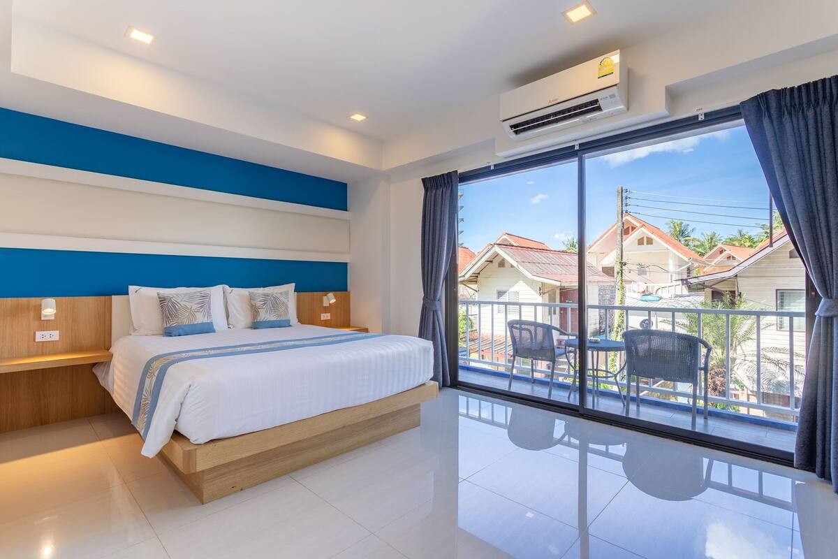 Ein modernes Hotelzimmer mit einem großen Bett, einem Balkon mit Sitzgelegenheiten und Blick auf angrenzende Häuser.