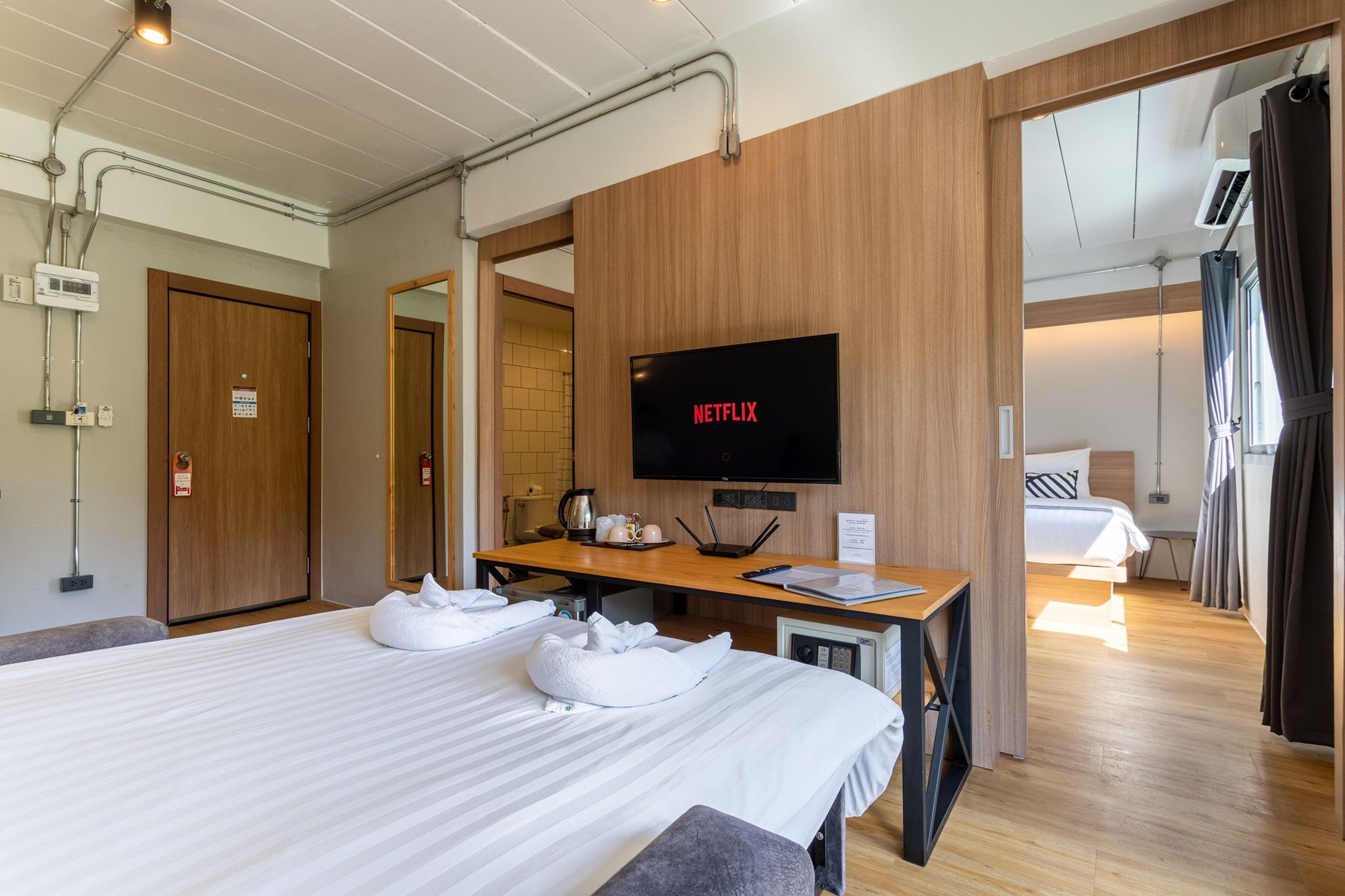 A hotel room with a large bed, a wooden desk, a television displaying 'NETFLIX', and a separate bedroom with a bed and curtains.