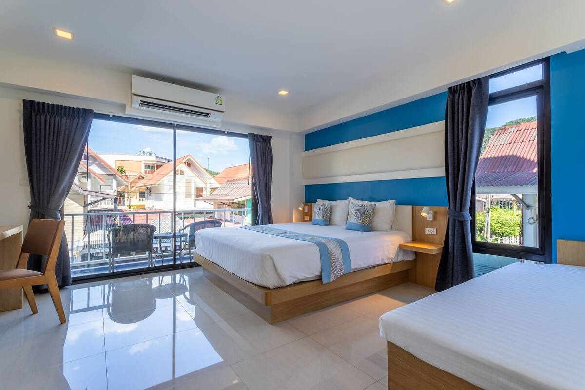 Ein Hotelzimmer mit einem großen Bett, einem Einzelbett, einem Sessel, einem Tisch und einem Balkon mit Blick auf Gebäude.