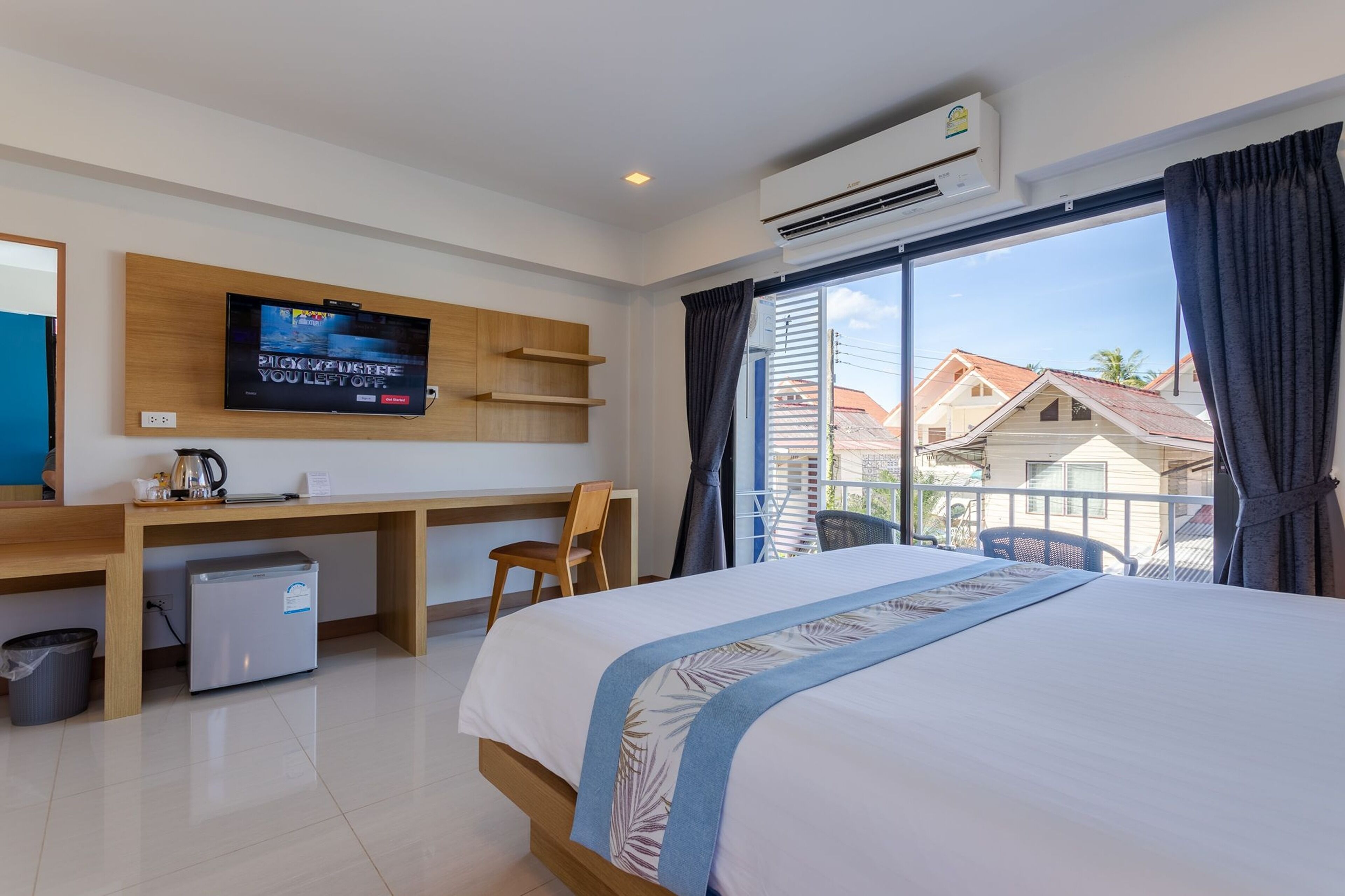 Habitación de hotel con cama, escritorio con televisor, refrigerador pequeño y vista a casas por la ventana.