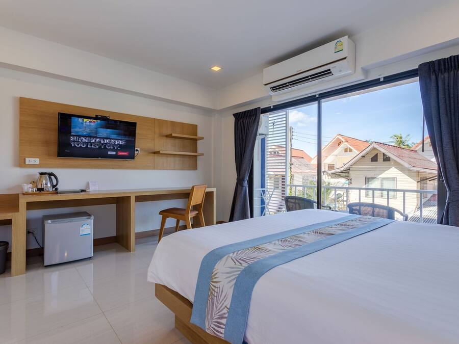 Habitación de hotel con cama, escritorio con televisor, refrigerador pequeño y vista a casas por la ventana.