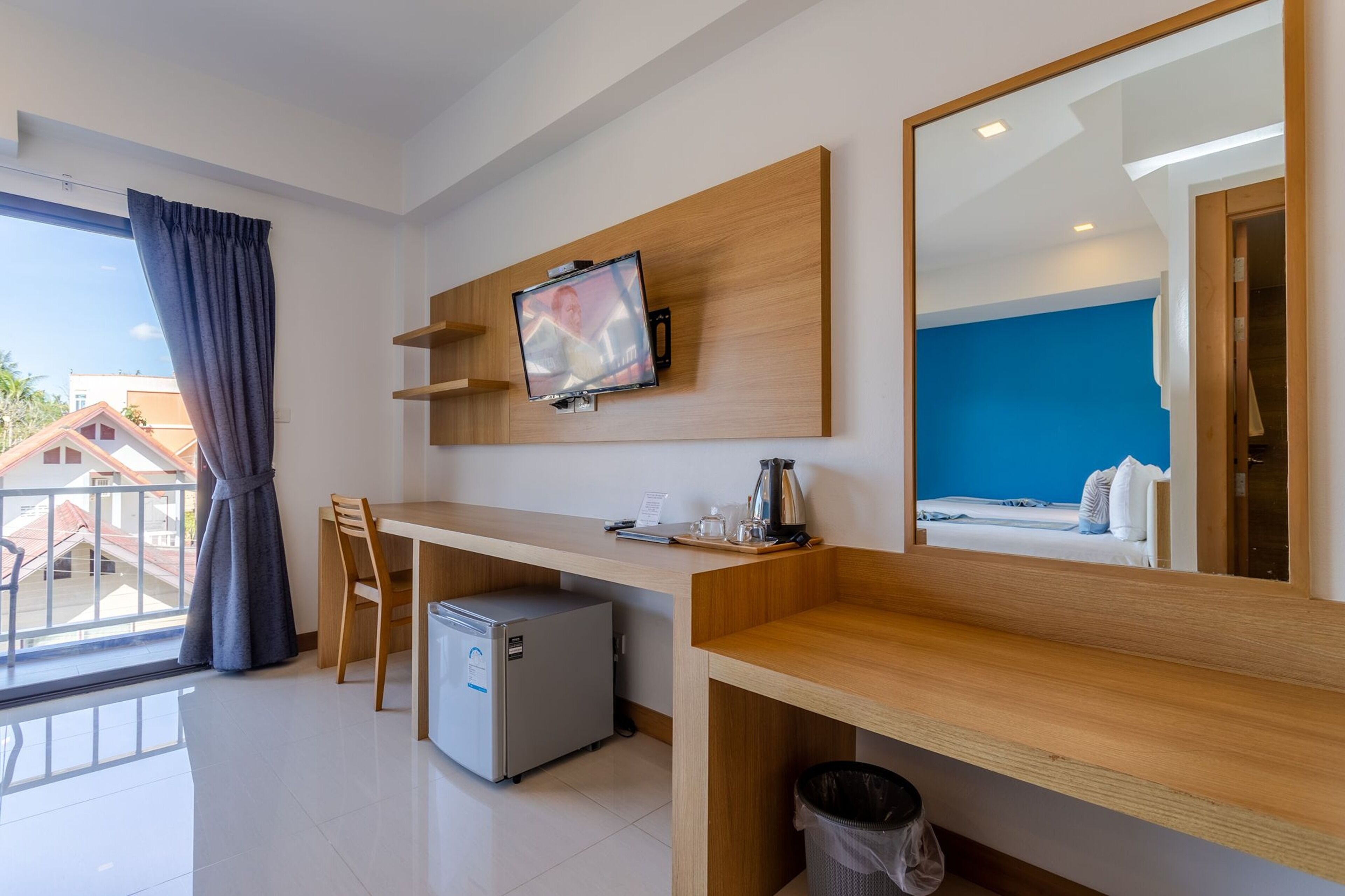 A hotel room with a wooden desk, a television mounted on the wall, a small refrigerator, and a bed with a blue wall.