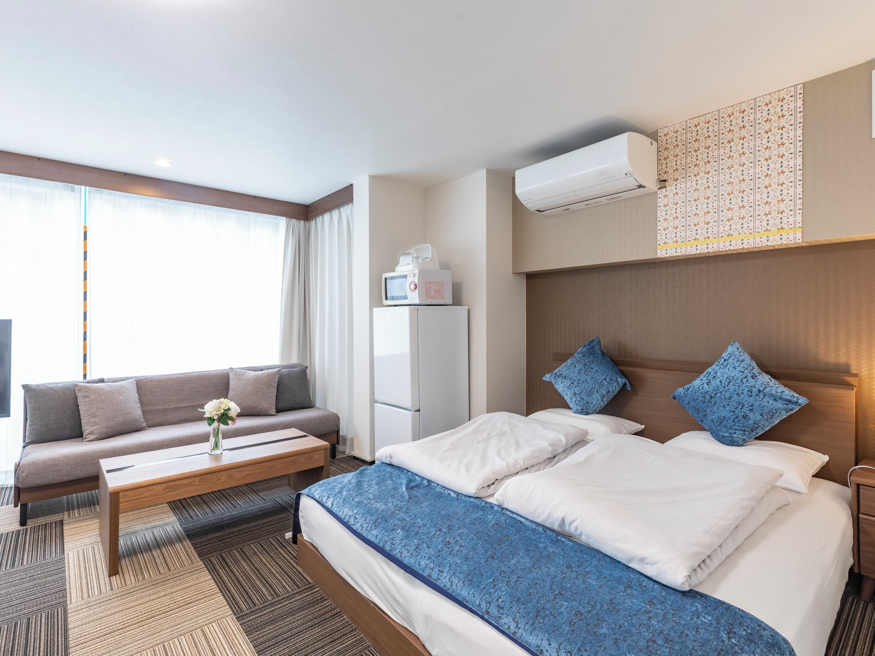 A hotel room with a bed, a sofa, a coffee table, a microwave, and an air conditioning unit.