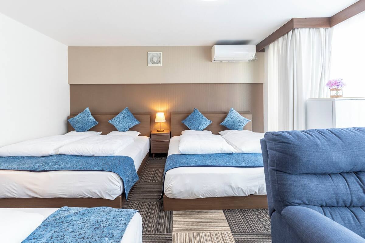 A hotel room with two beds, a nightstand with a lamp, an air conditioning unit, and a window with curtains.