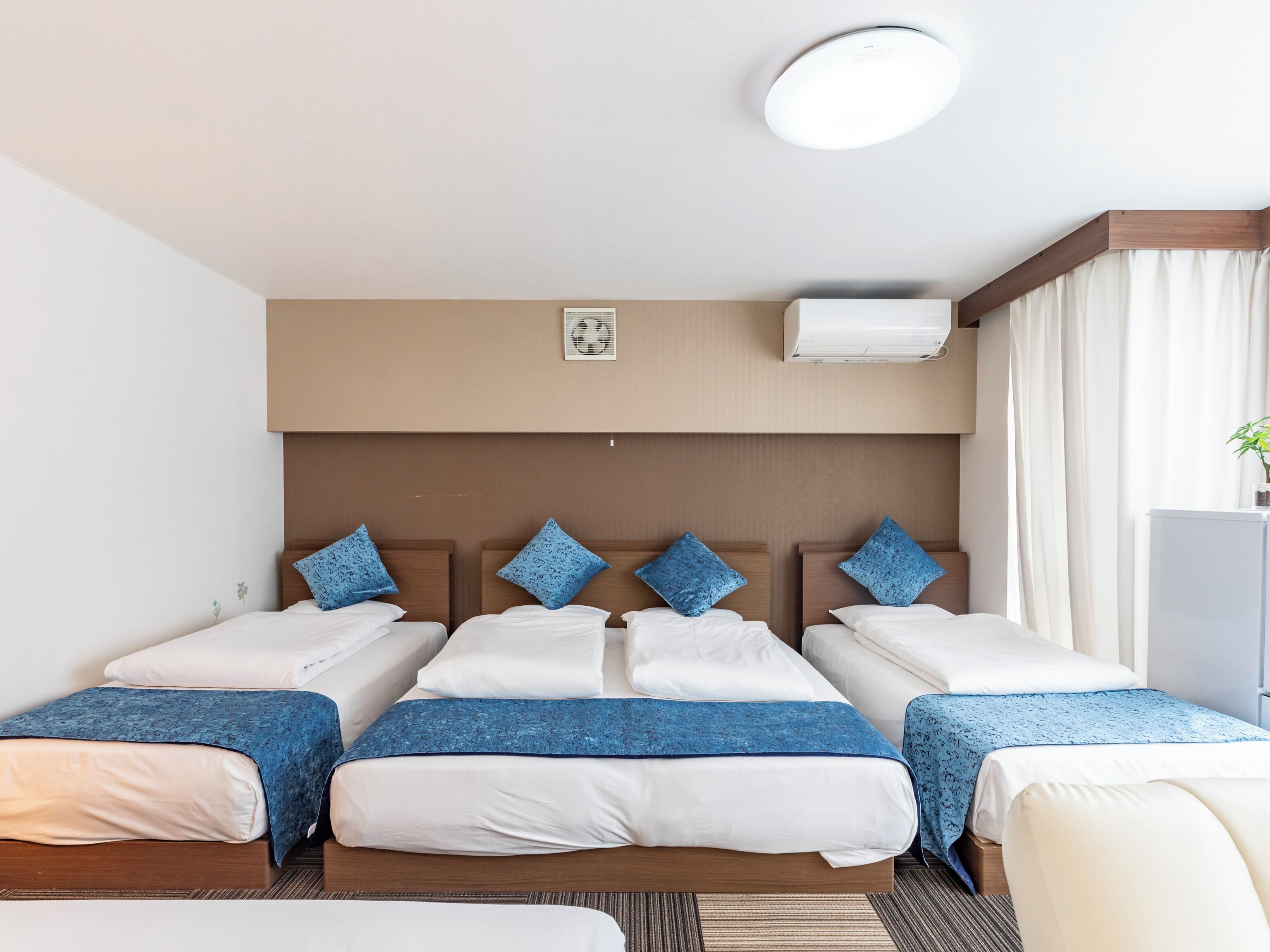 Een hotelkamer met drie bedden, een plafondventilator, een airconditioner en een raam met gordijnen.