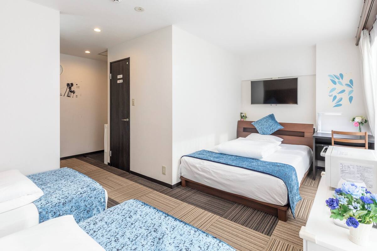 A hotel room with a bed, two bedside tables, a desk with a chair, a TV mounted on the wall, and a door leading to another room.