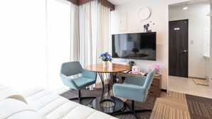 A modern hotel room with a large flat-screen TV, two blue armchairs, a white sofa, a round dining table with a vase of flowers, and a wooden door labeled "TOILET". - Tabist Sakuragawa River Side Hotel (Osaka)