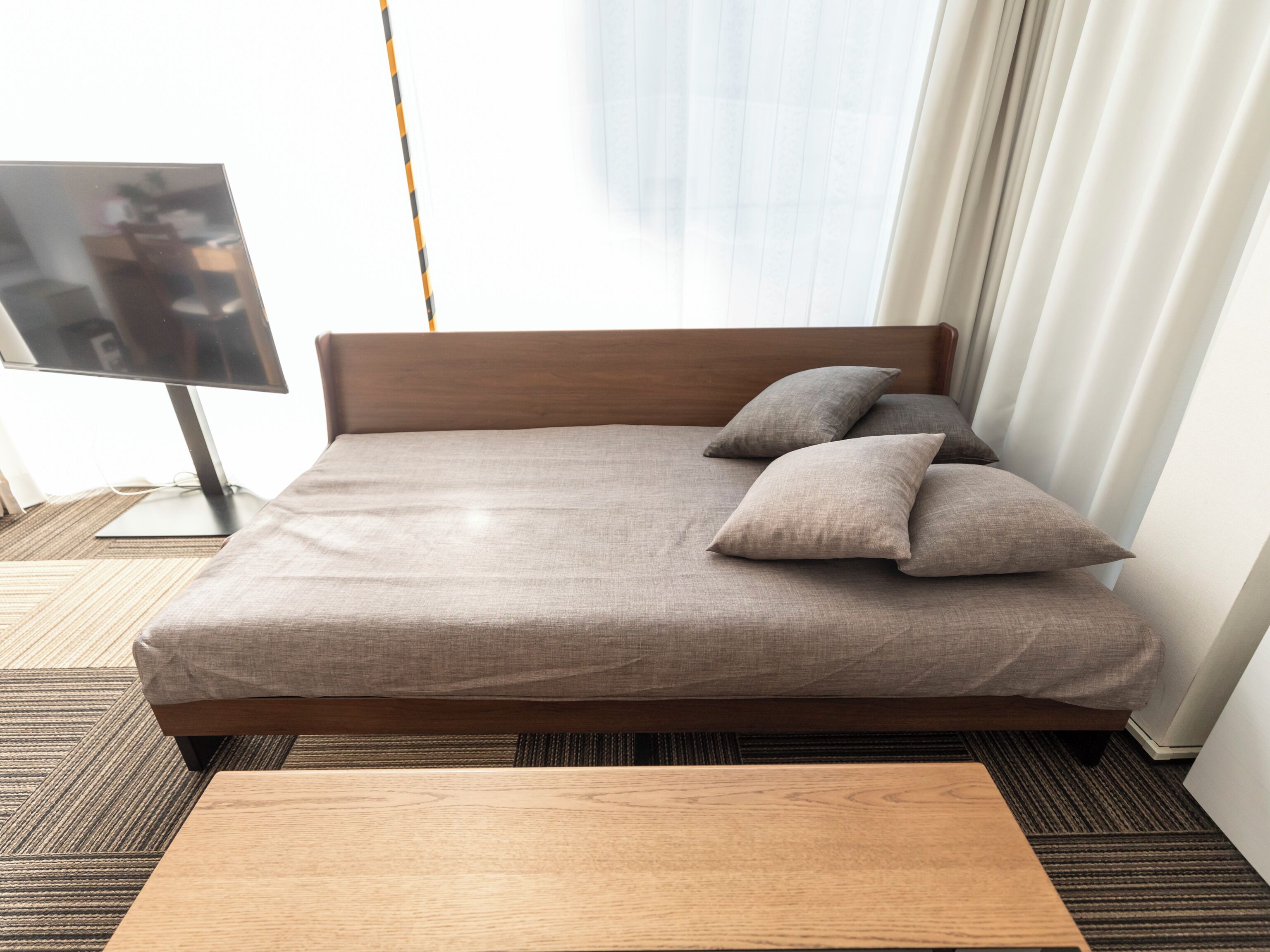 A neatly made bed with a wooden headboard, a small wooden table in front, and a television on a stand to the left.