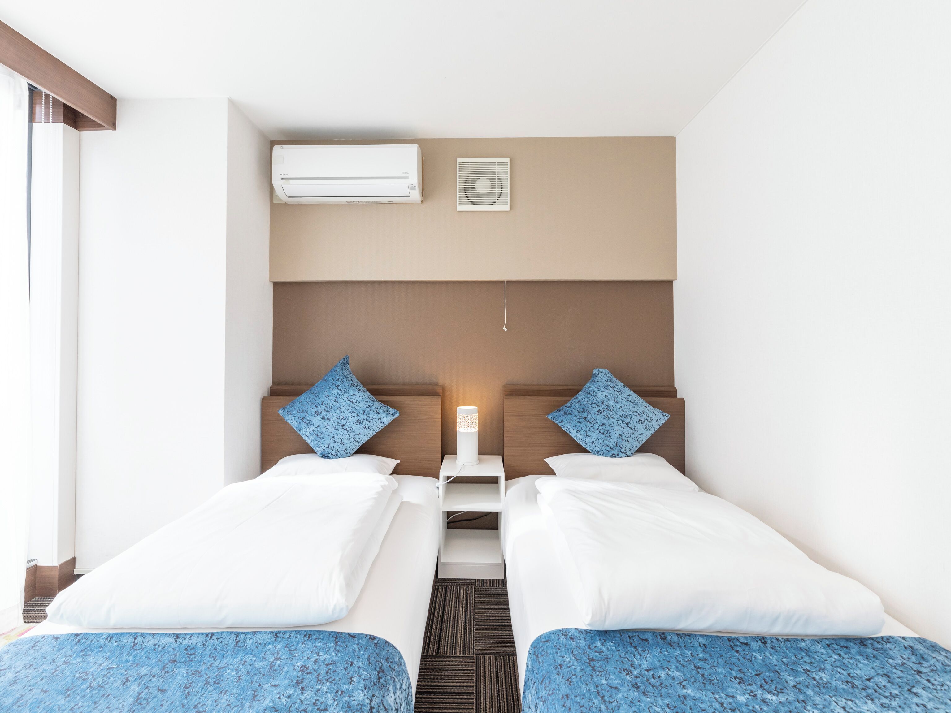 Une chambre d’hôtel avec deux lits, une table de chevet, un appareil de climatisation, une fenêtre et un miroir fixé au mur.