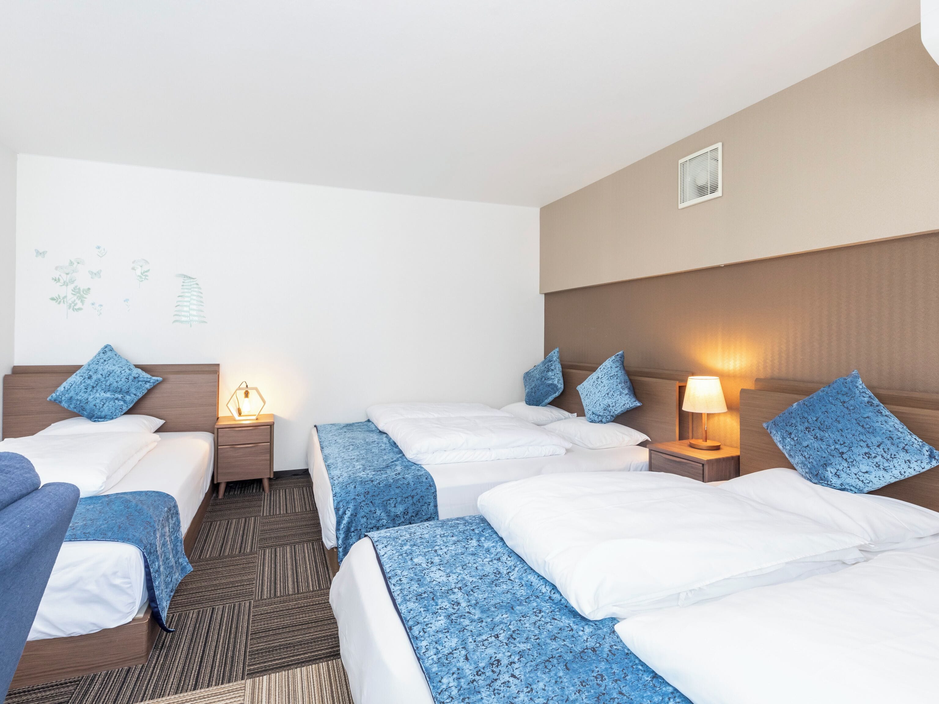 A hotel room with two beds, each with a blue floral bedspread, a nightstand with a lamp, and a wall-mounted heater.