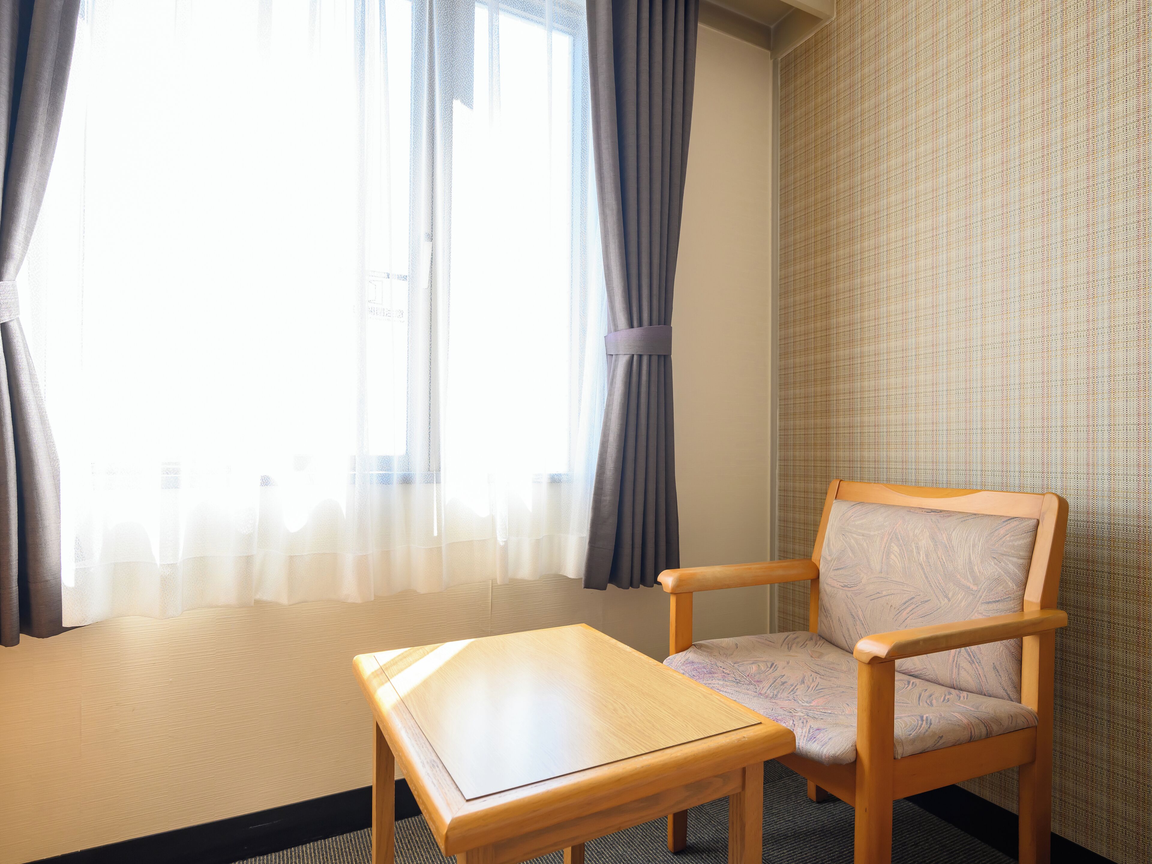 Una habitación con una silla de madera y una pequeña mesa, una ventana con cortinas, y una pared con diseño.