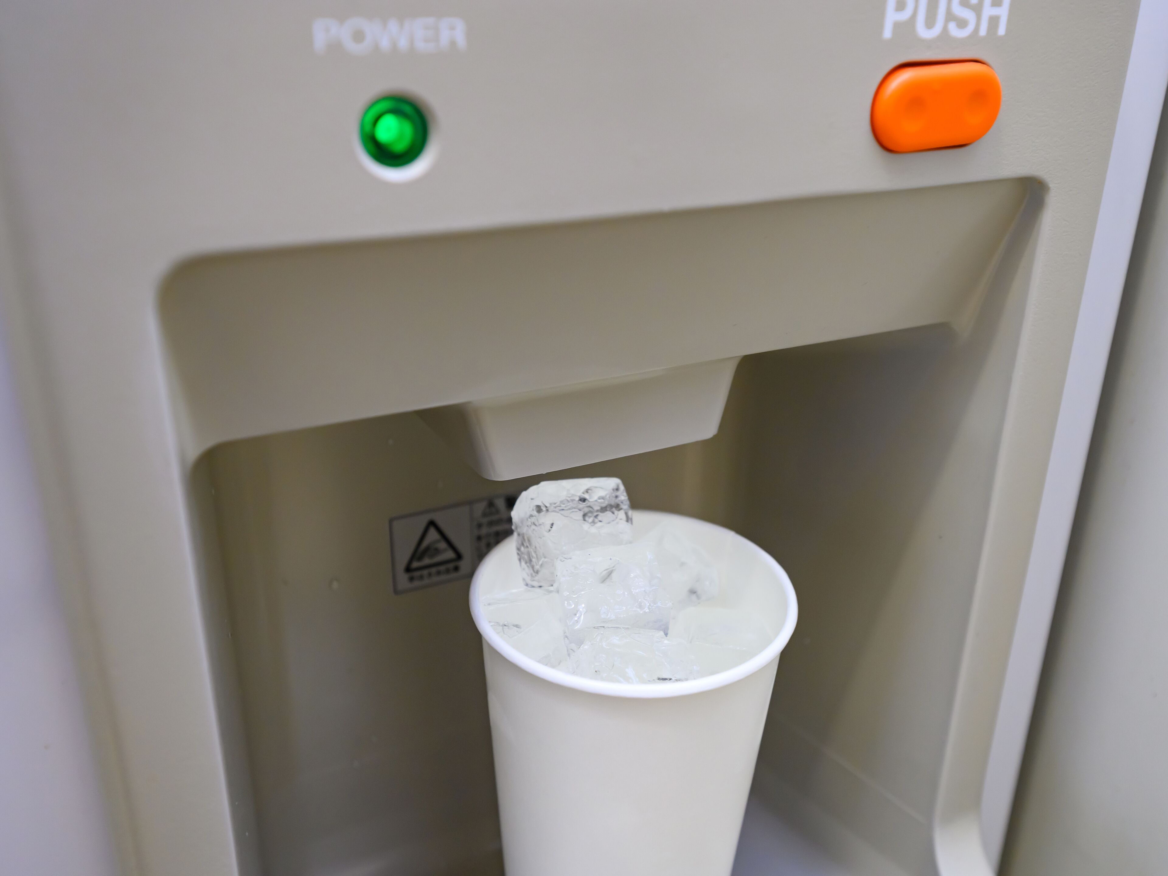 A water dispenser with a green power button and an orange push button, dispensing ice into a cup.