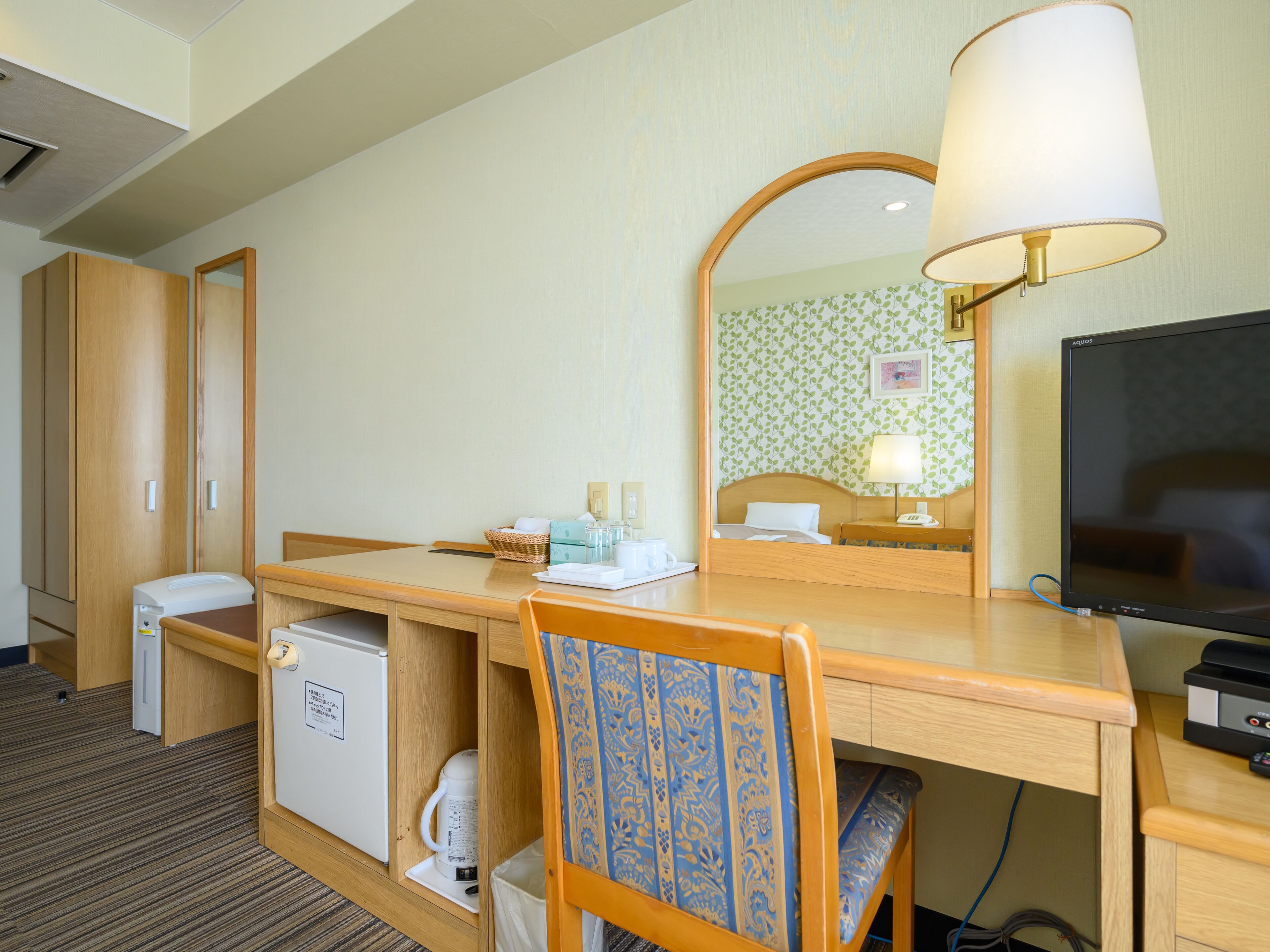 Ein Hotelzimmer mit Holztisch, Stuhl, Fernseher, Spiegel, Bett, kleinem Kühlschrank und Nachttisch mit Lampe.