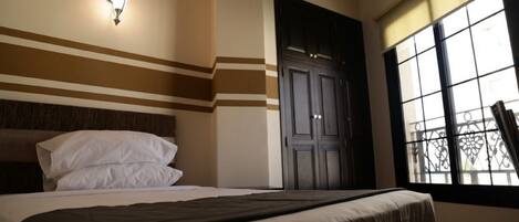 A hotel room with a bed, a wall-mounted light fixture, a window with a view, and a door.