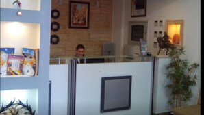 A reception area with a receptionist behind a glass partition, a framed picture on the wall, and a potted plant. - Fantazia Hotel (Oran)