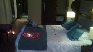 Une chambre d’hôtel avec un lit, des oreillers, une table de chevet avec une lampe et une petite table ronde sur laquelle sont disposés des fruits et des bougies.