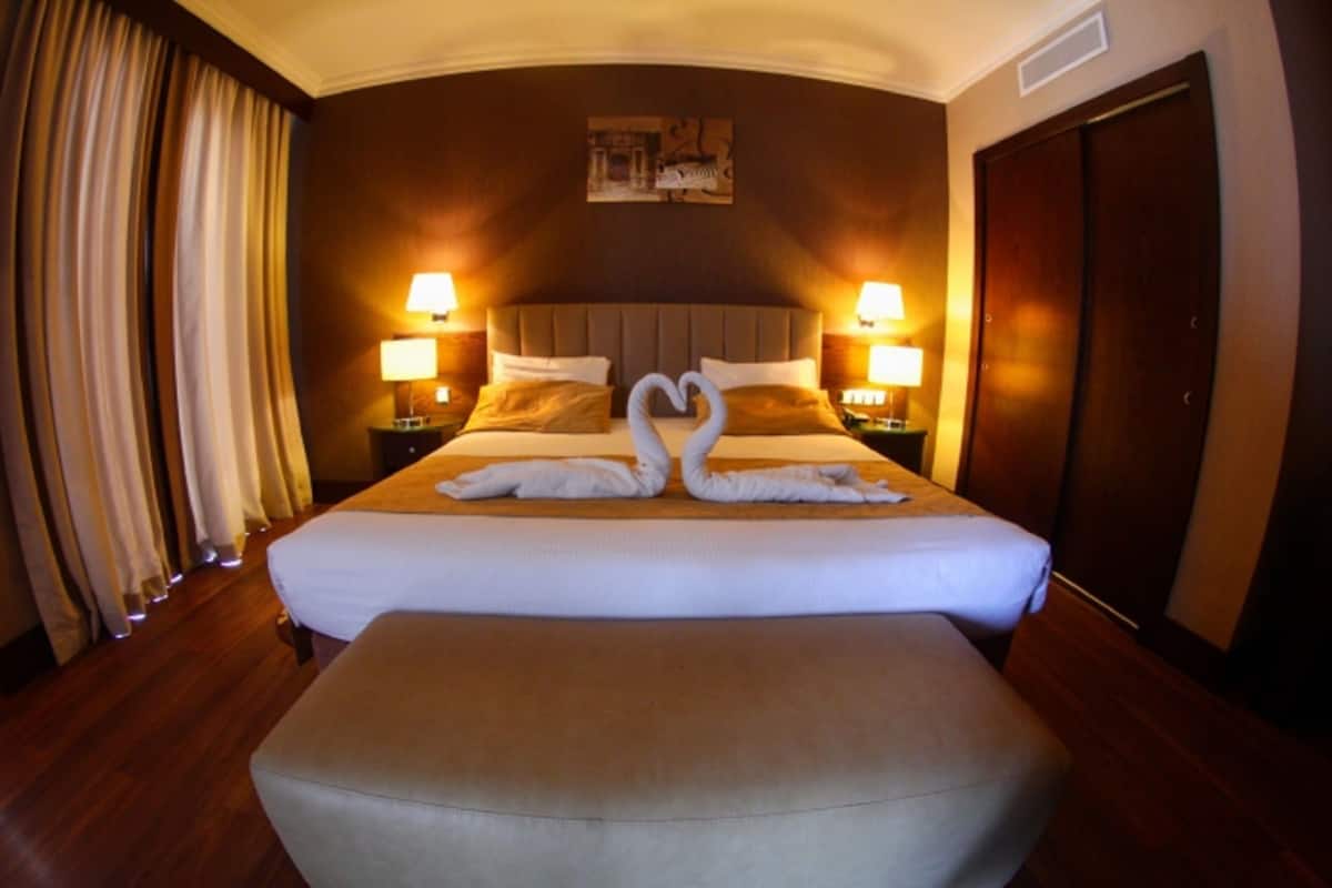 A hotel room with a large bed, two bedside lamps, a headboard, a painting on the wall, and a wooden floor.