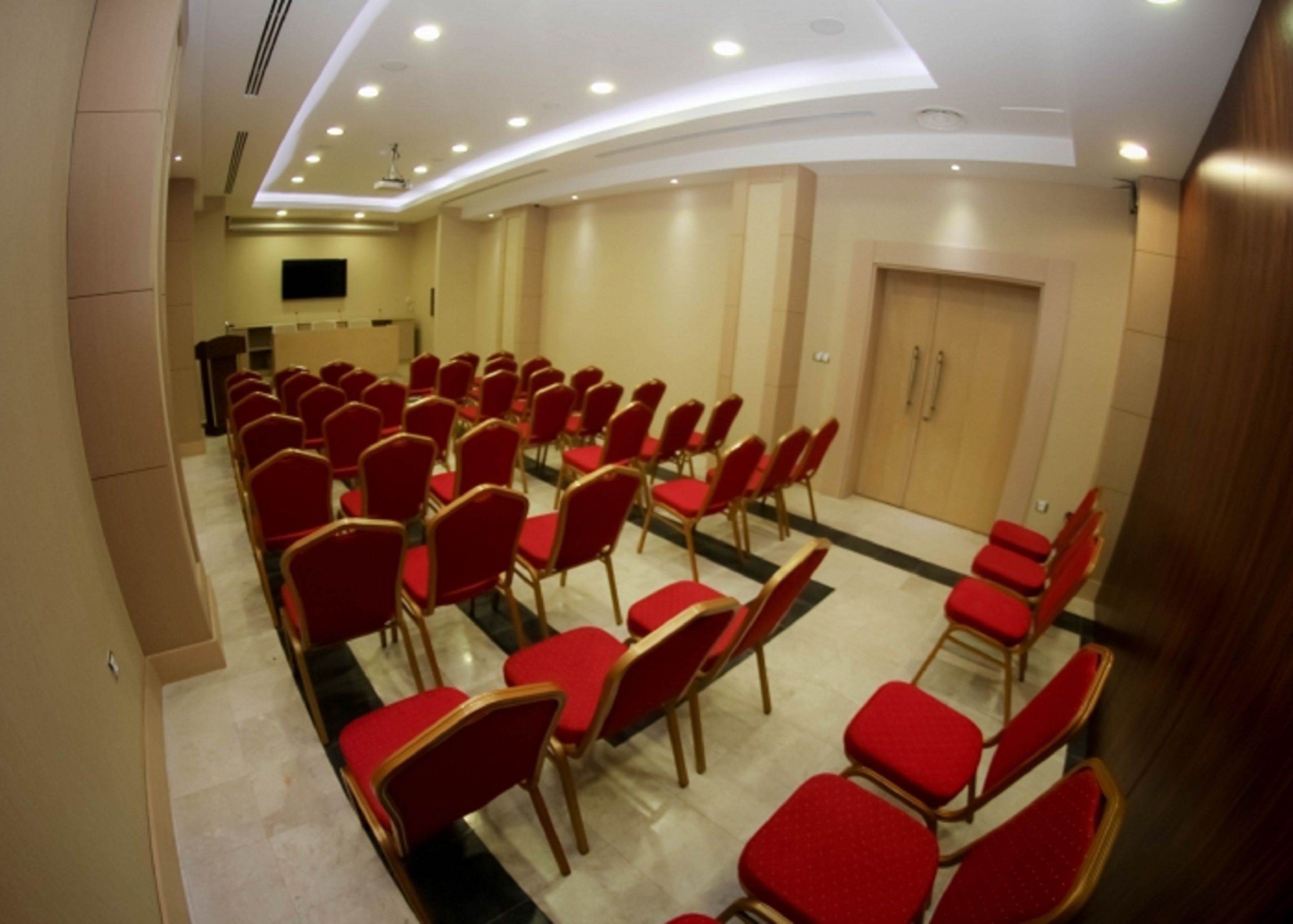 A conference room with rows of red chairs, a podium, and a flat-screen TV on the wall.