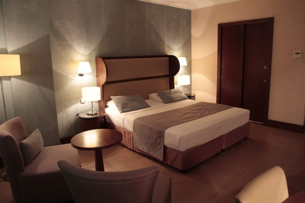 A hotel room with a large bed, two bedside tables with lamps, a round coffee table, and two armchairs.
