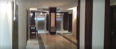 En moderne hotelgang med en central elevator, afgrænset af døre på begge sider.