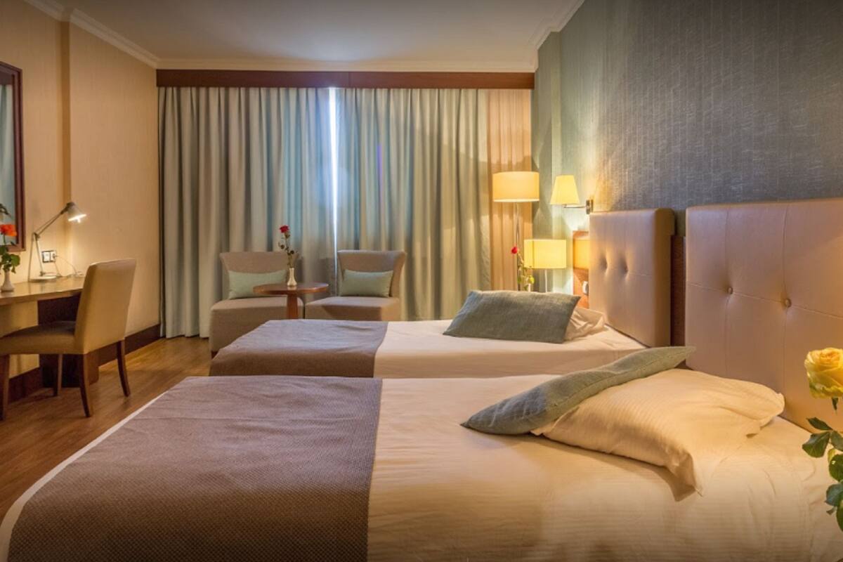 A hotel room with two beds, a desk, chairs, and a large window with curtains.