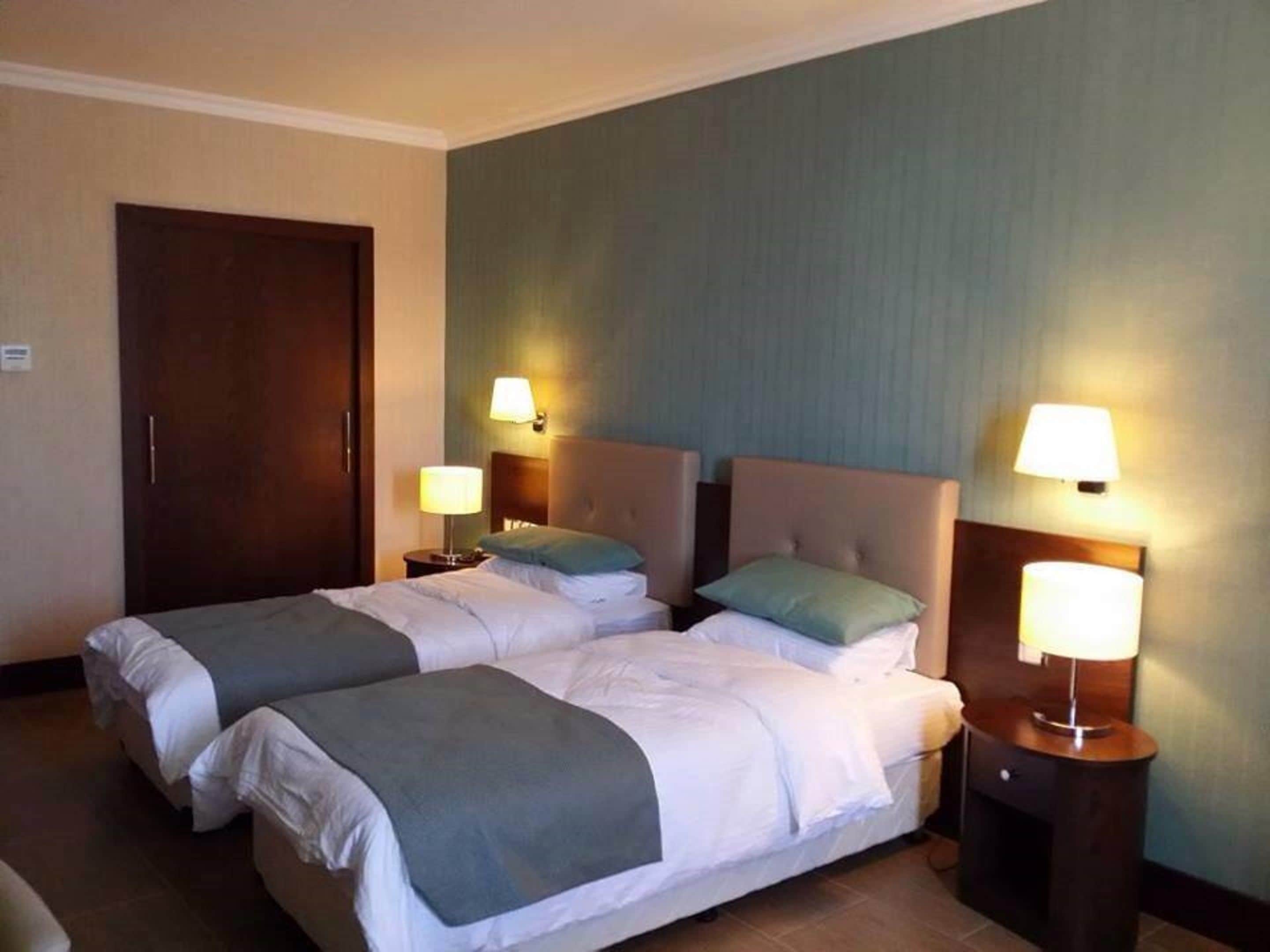A hotel room with two beds, each with a headboard, bedside tables with lamps, and a door.