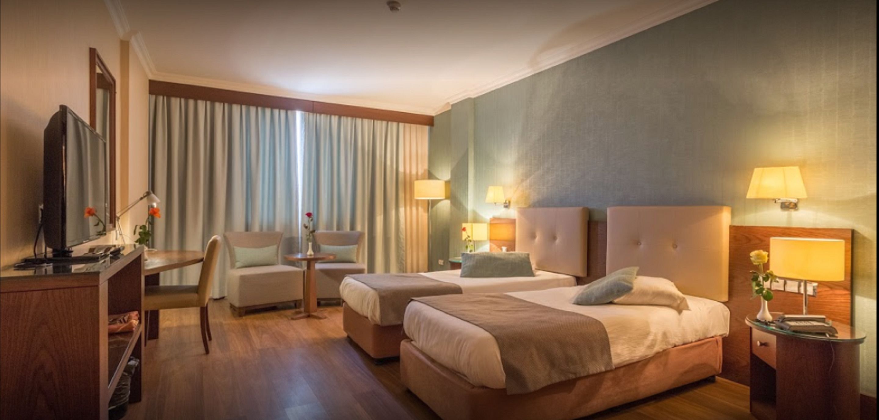 A hotel room with two beds, a desk, a chair, a TV, and lamps.