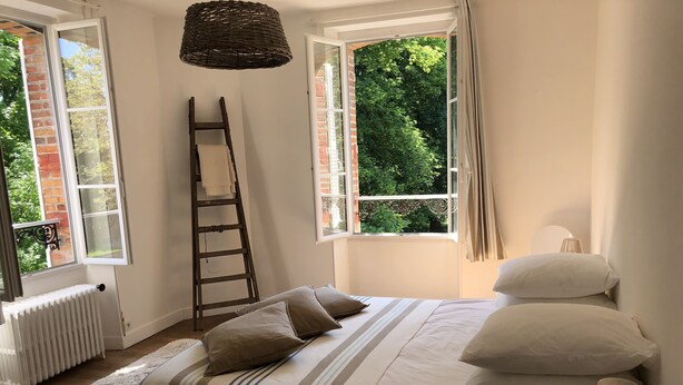 Un dormitorio con cama, almohadas, una escalera, un radiador y una ventana con vistas a vegetación.