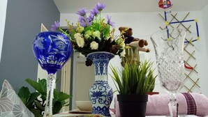 Une table avec un vase bleu, un vase blanc avec des fleurs violettes, un ours en peluche, une plante en pot et un verre transparent.