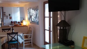 Un comedor con una mesa y sillas de vidrio, un televisor montado en la pared y una cocina al fondo.