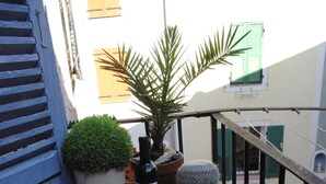 Una pequeña mesa al aire libre con una botella de vino, una copa y un cuenco de fruta. Hay una maceta y un cactus sobre la mesa. Un cojín a rayas y de colores descansa sobre una silla de mimbre junto a la mesa.