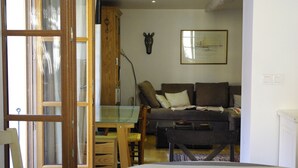 Un comedor con una mesa dispuesta para dos, un frutero y vista a una sala de estar con un sofá y un cuadro en la pared.