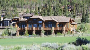 A large log cabin with multiple balconies and a stone foundation, surrounded by a fenced property and a grassy field. - Five Star Grand Lodge @ Peak 7:  Week of July 4th! (Breckenridge)