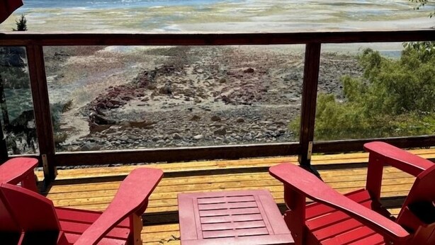 Eine Holzterrasse mit roten Stühlen und einem Tisch, mit Blick auf eine felsige Küstenlinie und ein ruhiges Meer mit Bergen in der Ferne.